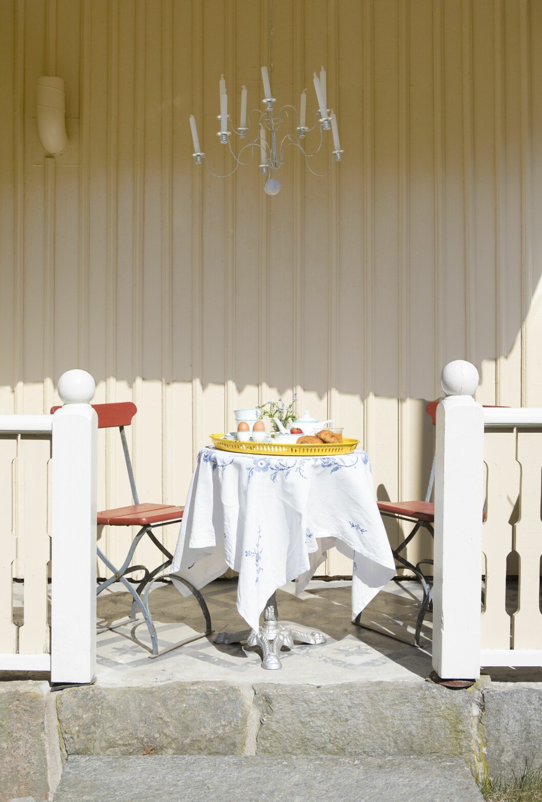 Att äta frukost ute en skön sommardag slår det mesta. Duka vackert med en broderad duk som du köper på loppis för några tior. Veranda med bord och två stolar. På bordet ligger vit duk med blått mönster och en gul bricka dukad med ägg, croassianter och vit teservis.