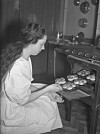Lucia 1939 i Sverige. En ung kvinna i luciadräkt bakar lussebullar/lussekatter och sätter in en plåt i ugnen. Köket har en gasugn och det hänger härligt nostalgiska kopparformar i bakgrunden.
