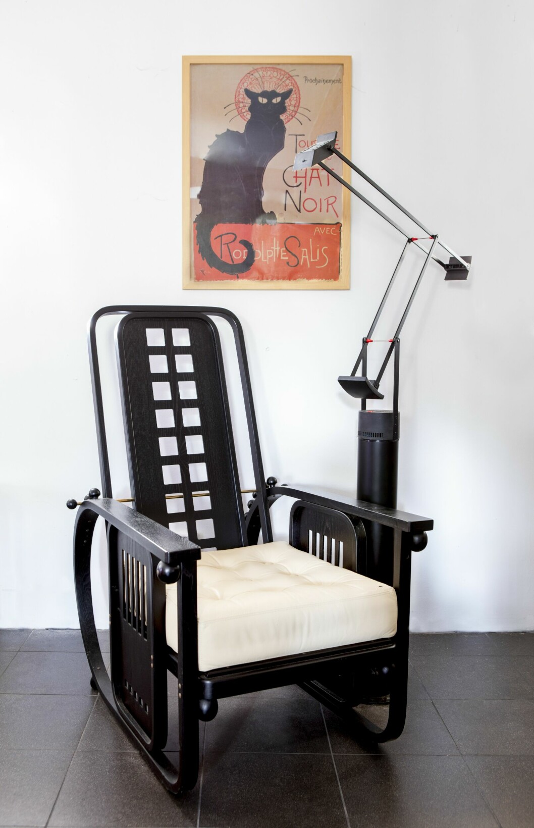 Sittmaskiner brukade Josef Hoffmann kalla sina stolar och fåtöljer. Här har vi en reproduktion, tillverkad i Italien på 1970-talet då en ny jugendvåg svepte över världen. Originalmodellen formgavs 1908 och tillverkades i färgat, laminerat och böjt bokträ. Med hjälp av kulorna, en detalj som ofta återkommer på Hoffmanns möbler, kan ryggen justeras i olika lägen. Vilstolen finns hos Palk Antik för 5 000 kr. Svart vilstol med vit dyna, tavla med katt på väggen och svart stålampa.