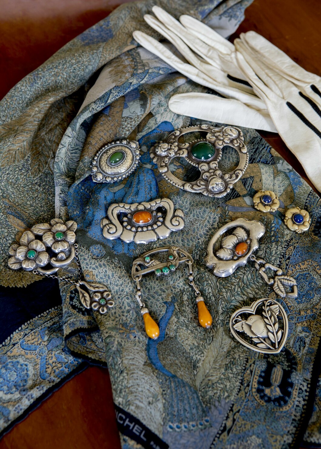 Silversmycken från förra sekelskiftet. Broschen med små agater och droppformade bärnstenshängen är stämplad Georg Jensen, liksom den hjärtformade broschen med fågel. Silverknapparna med sten av lapis lazuli är stämplade Evald Nielsen. Broschen längst till vänster med krysopraser och silverkedja kommer från den norske guldsmeden David Andersen. Den stora broschen med agat i mitten är stämplad Nicklas Muck. Övriga smycken är av svensk anonym design. Allt från Palk Antik, där priserna varierar mellan 2 500–5 000 kr. Smycken med färgglada stenar på en scarf och vita handskar.