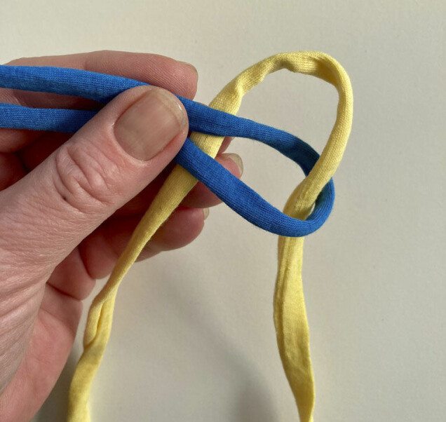 Trä in det gula bandet underifrån genom den blå öglan och ner bakom. Trä in det gula bandet underifrån genom den blå öglan och ner bakom.
