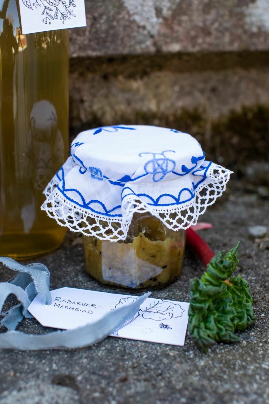 En burk rabarbermarmelad ser extra festlig ut med en liten duk på locket. Bara att lägga över en rund duk och knyta ett snöre runt. Glasburk med rabarbermarmelad med en blåvitmönstrad duk som lock. Handskriven etikett ligger framför.