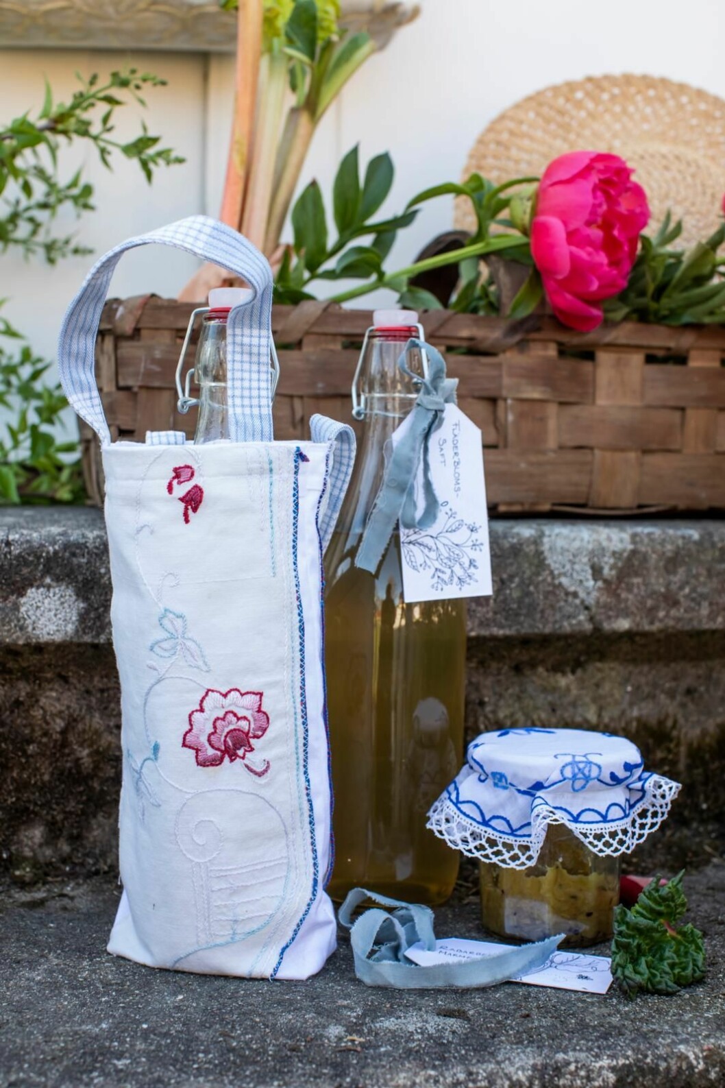 Bästa gå bort-presenten i sommar – en handsydd flaskväska med en flaska fläderblomssaft i. Utgå från grundmönstret till tygkassen, men använd måtten 18x30 cm till väskan och måtten 6x35 cm till hantagen. Två saftflaskor i glas, den ena nedstoppad i en vit- och blåmönstrad tygkasse, i bakgrunden syns en kort med en cerise pion i.