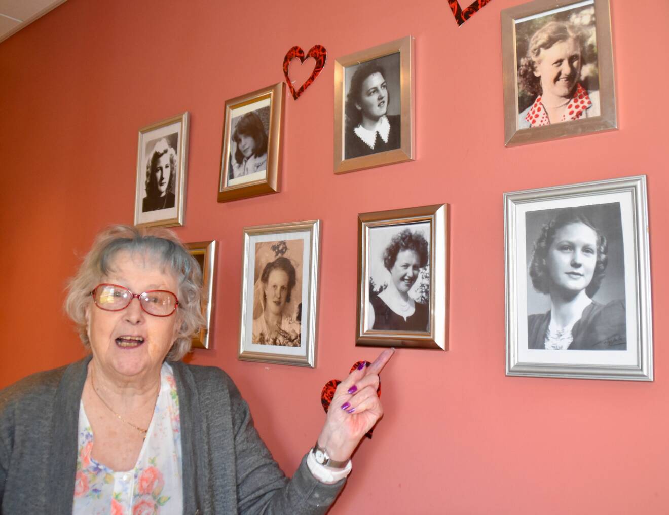 Äldre kvinna på äldreboendet står framför en vägg med bilder på de äldre som unga.