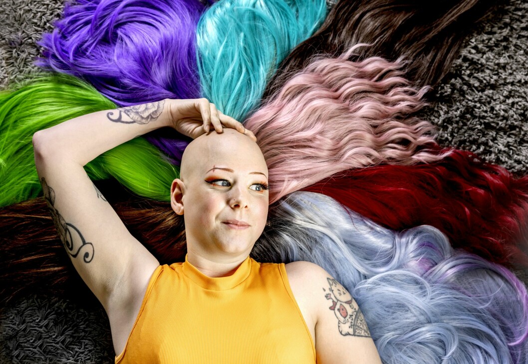 Lena Jakobsson, som har alopeci, ligger på rygg med sitt kala huvud vilande på en hög färgglada peruker. Hon har flera tatueringar och effektfullt sminkade ögon med orange ögonskugga.