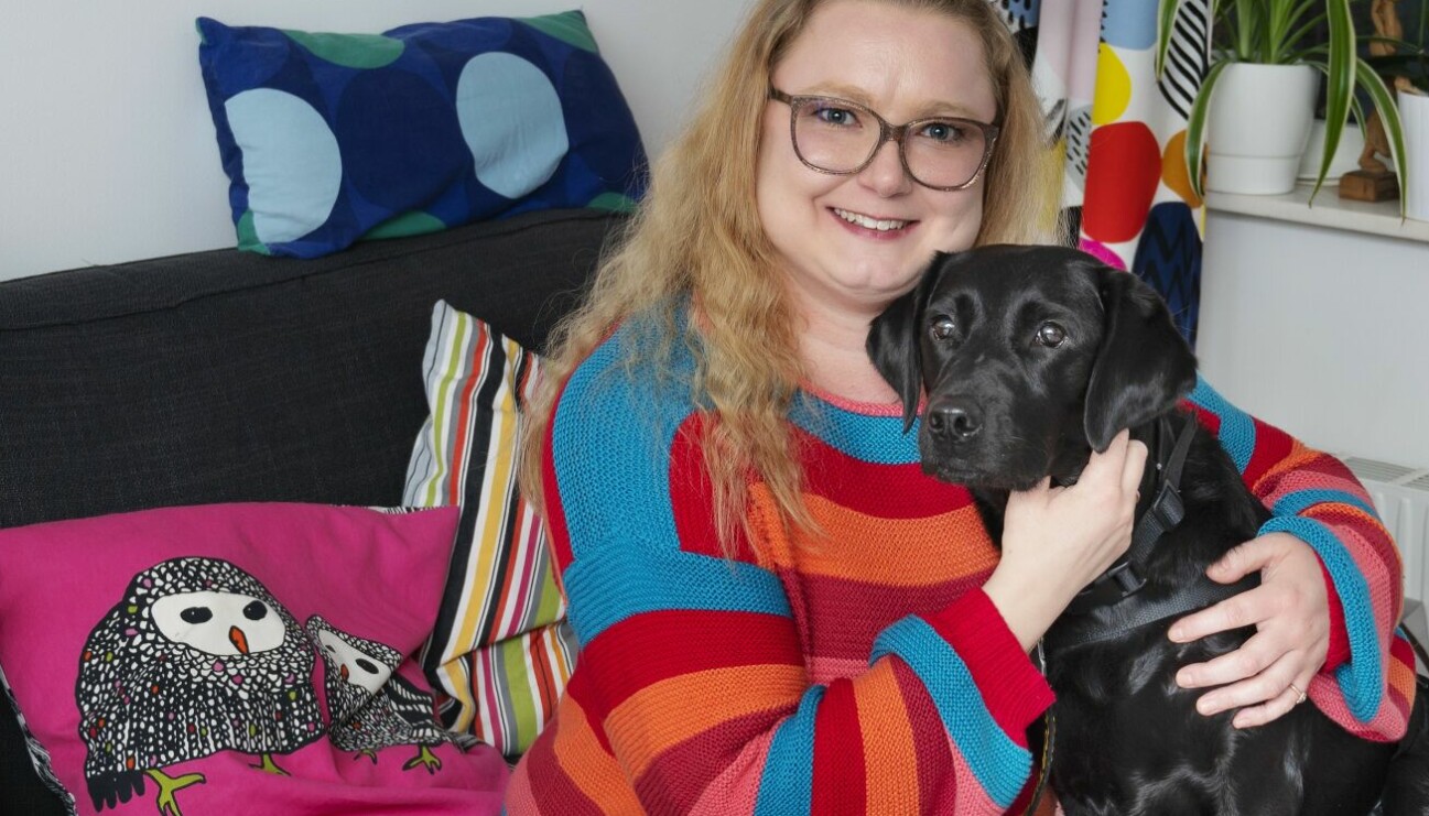 Anna-Maria sitter i soffan tillsammans med sin assistanshund Mini och berättar om hur det är att leva med sjukdomen binjurebarksvikt och hur hennes liv förändrades när hon skaffade assistanshund.