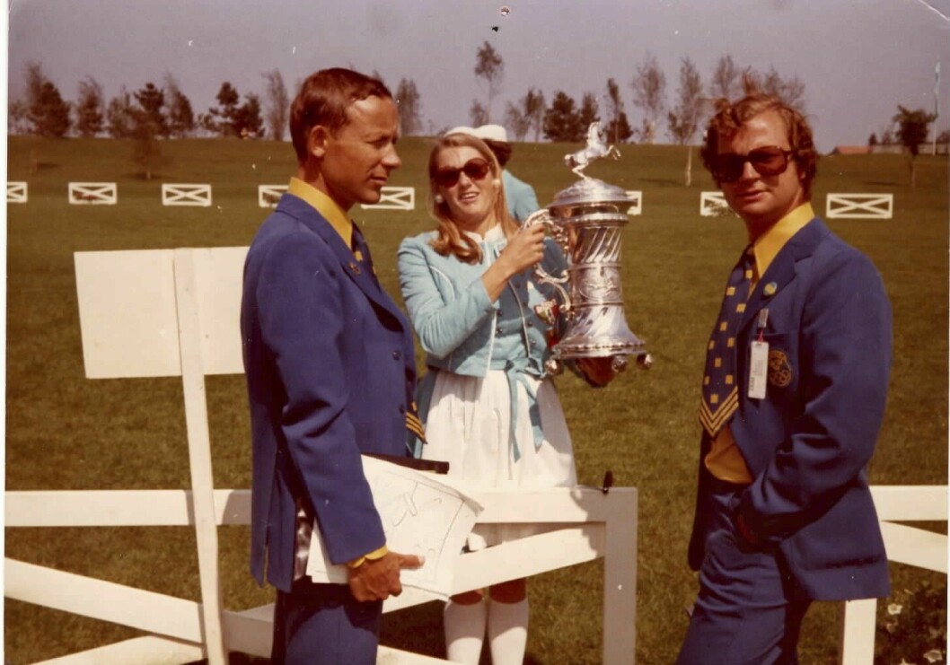 Den unga OS-värdinnan står och håller i någon form av ridsportpokal, där hon står vid ett fält med kronprins Carl Gustaf och hans adjutant.