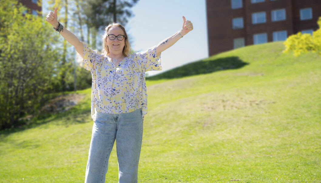 Arja Mattila står på en grön gräsmatta och tittar in i kameran samtidigt som hon gör tummen upp med båda händerna. Hon är klädd i ljusblå jeans och en mönstrad blus. I bakgrunden syns ett flervåningshus.