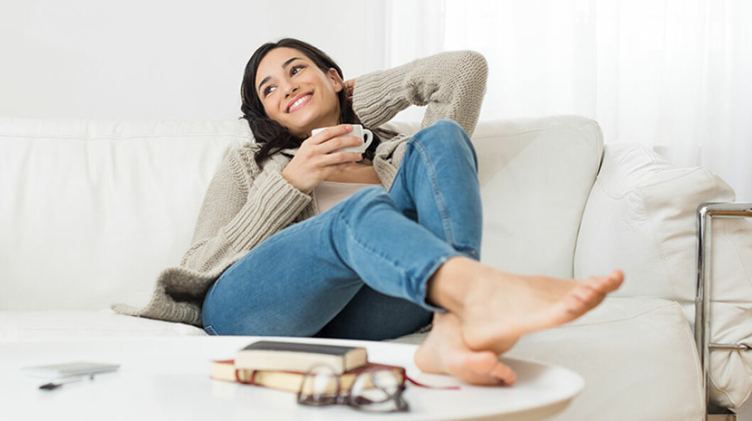 Bild på en kvinna som sitter i en ljus soffa och med fötterna på ett soffbord. Hon håller en kaffekopp i handen och ser glad ut.