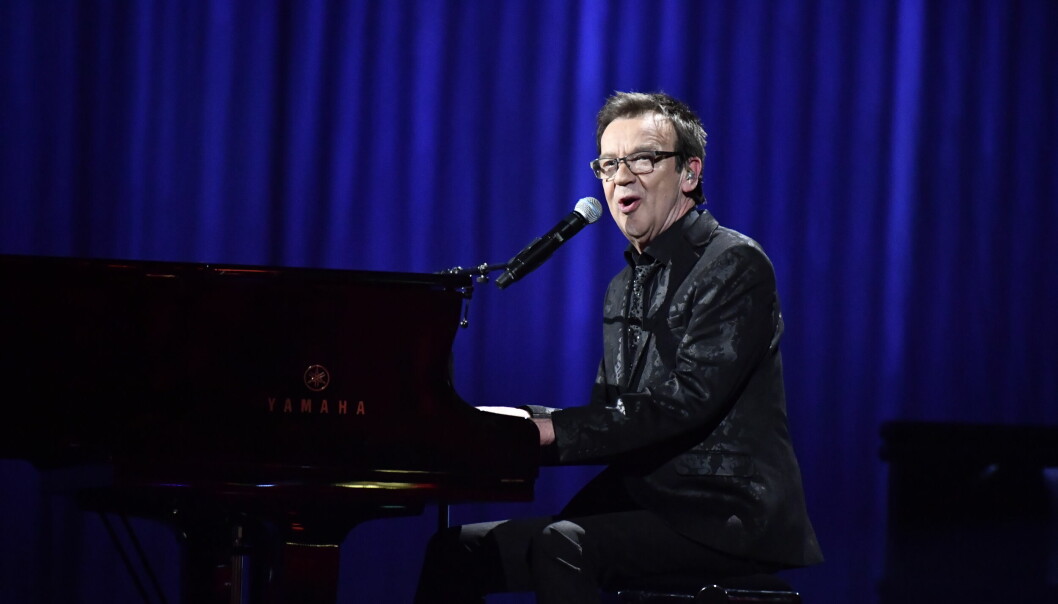Björn Skifs sitter vid ett piano som han spelar på och samtidigt sjunger i en mikrofon. Han är klädd i en glänsande svart kavaj.