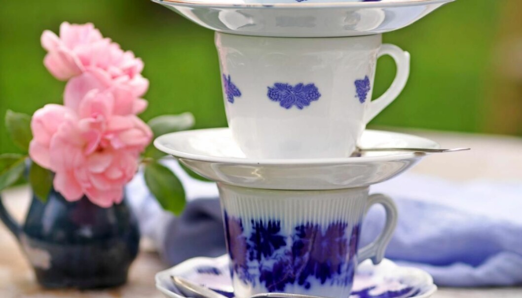 Tre blåvita kaffekoppar står staplade på varandra på ett träbord, bredvid två rosa blommor i en liten mörkblå vas.