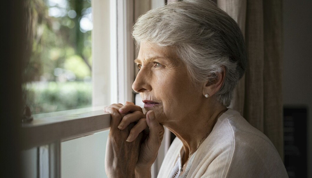 En äldre till synes deprimerad kvinna står och tittar ut genom ett fönster.