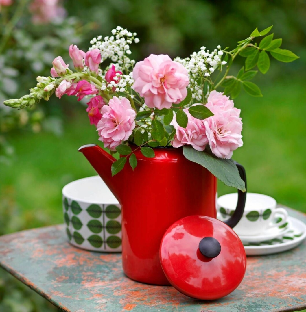 Emaljerad röd kanna med blommor står på ett bord tillsammans med delar ur Stig Lindbergs servis Berså. 