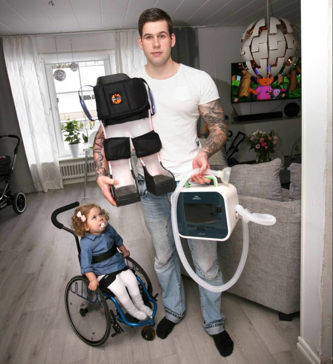 Stålskalet fixerar Ellys ben så att hon kan stå upp vid sitt lekbord och hostmaskinen hjälper henne att få upp slem som samlats i luftvägarna. Foto: Mikael Svensson Elly med sin utrustning såsom rullstol och hostmaskin.