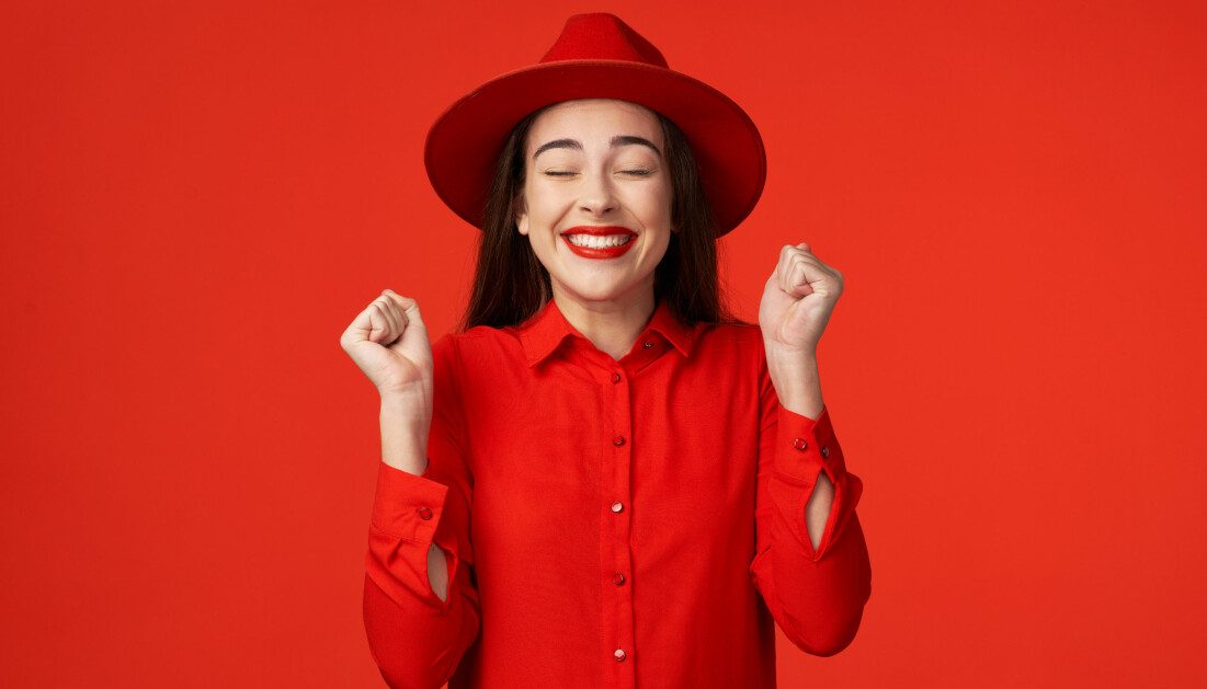 En kvinna i röd hatt, röd skjorta och rött läppstift står mot en röd bakgrund och ser glad ut.
