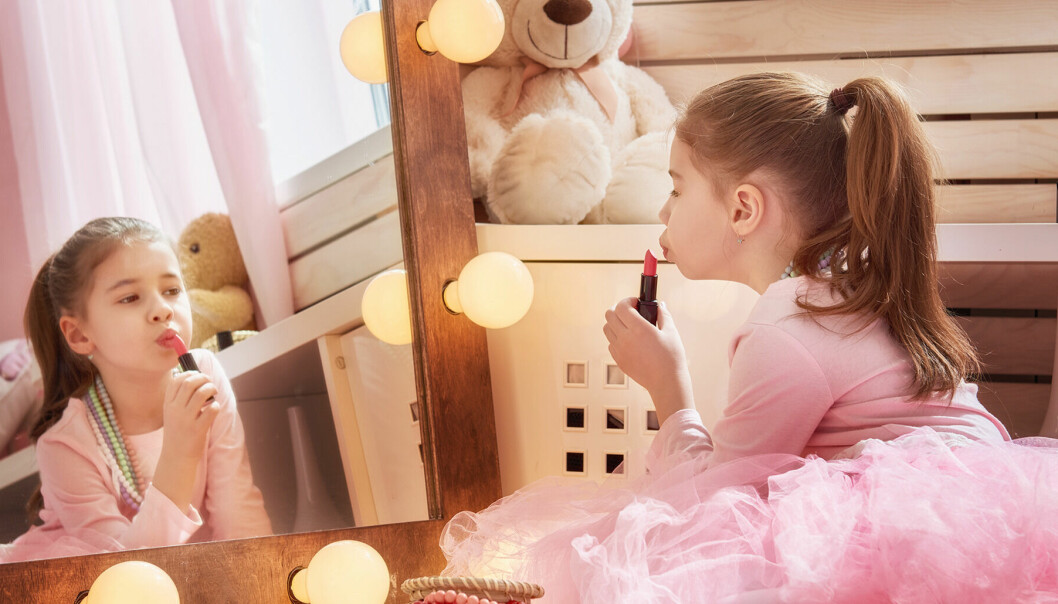 Brunhårig flicka i rosa ballerinaklänning sitter framför spegeln och målar läppstift på läpparna.