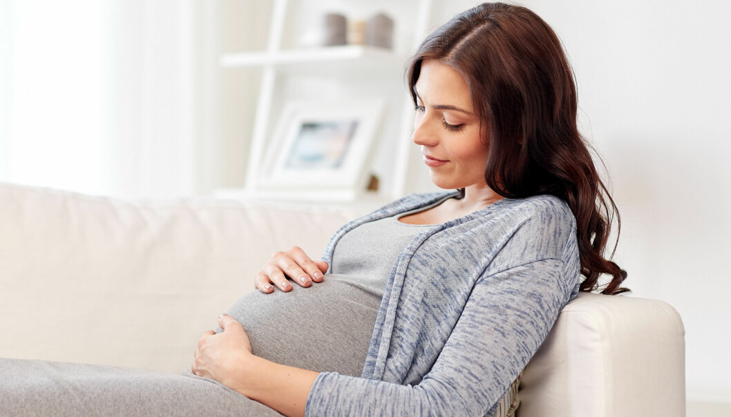 Gravid kvinna i 30-årsåldern sitter i en soffa och håller sina händer på sin mage.