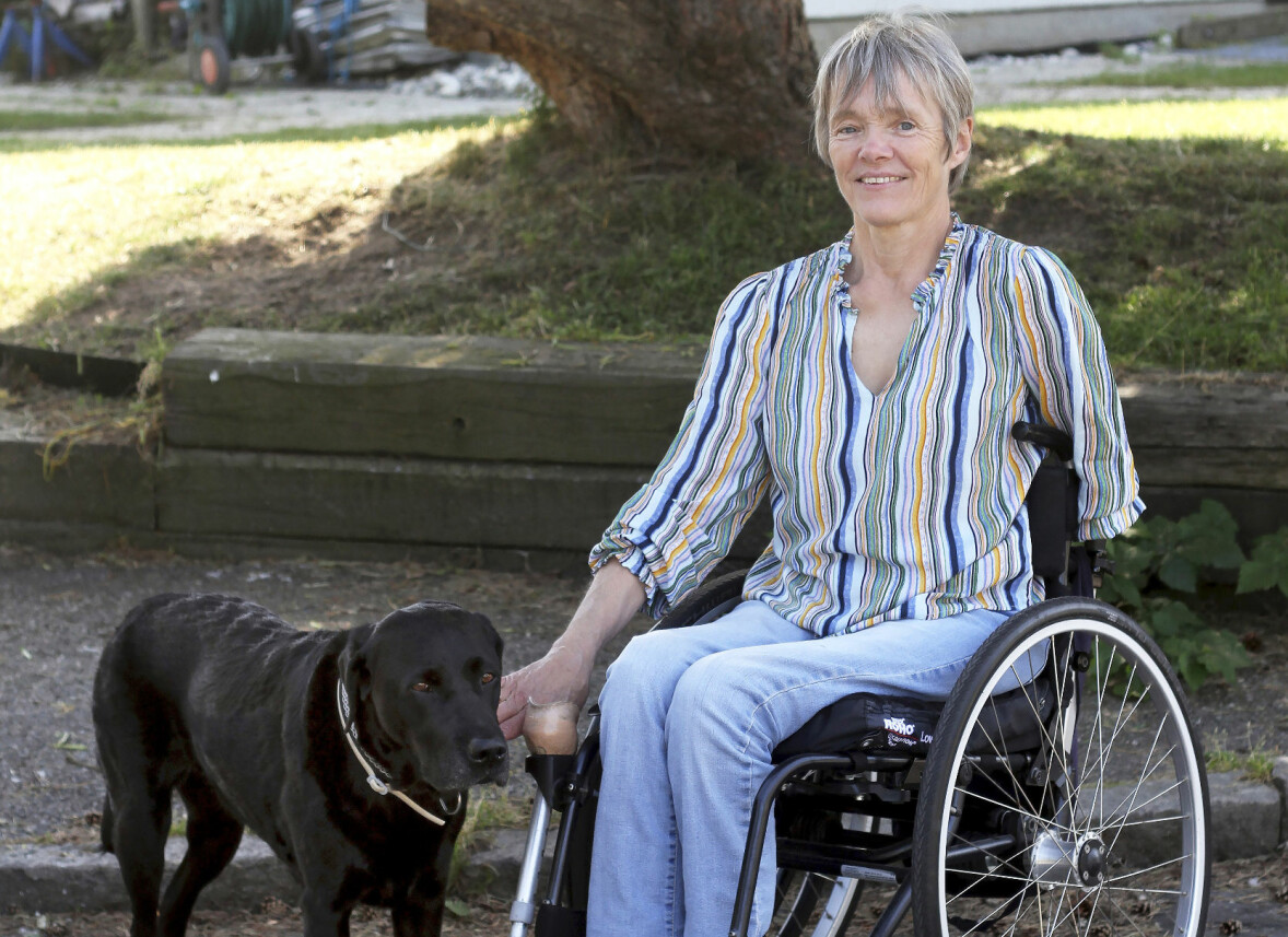 Gunilla sitter i sin rullstol utomhus framför ett träd och har den svarta labradoren och servicehunden Sassa bredvid sig