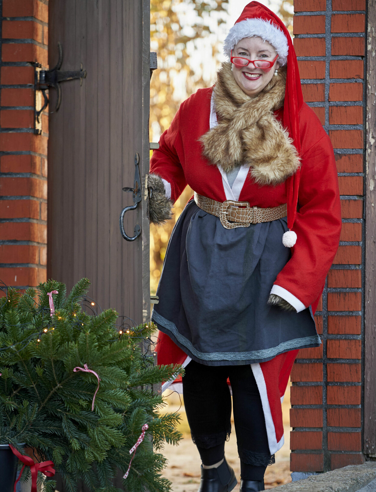 ”Jag är alldeles för kvinnlig för att kunna klä mig som en jultomte”, säger Ulrika om valet att i stället vara Tomtemor. Foto: Stefan Lindblom Ulrika, utklädd till Tomtemor, kikar leende fram genom en trädörr.