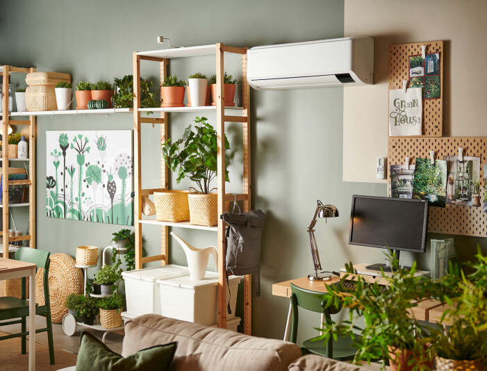 En vägg som är grön- och beigemålad. Längs väggen står en rymlig öppen hylla full av växter och förvaring. Till höger syns ett skrivbord med dator och anslagstavlor ovanför. På väggen hänger en luftvärmepump.