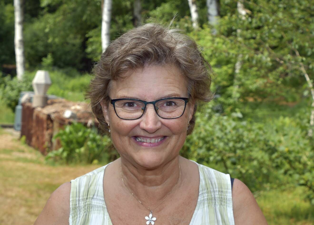 Susanne Karlsson står i trädgården, ler. Hon bär glasögon och ett halsband till ett grönvitt linne.