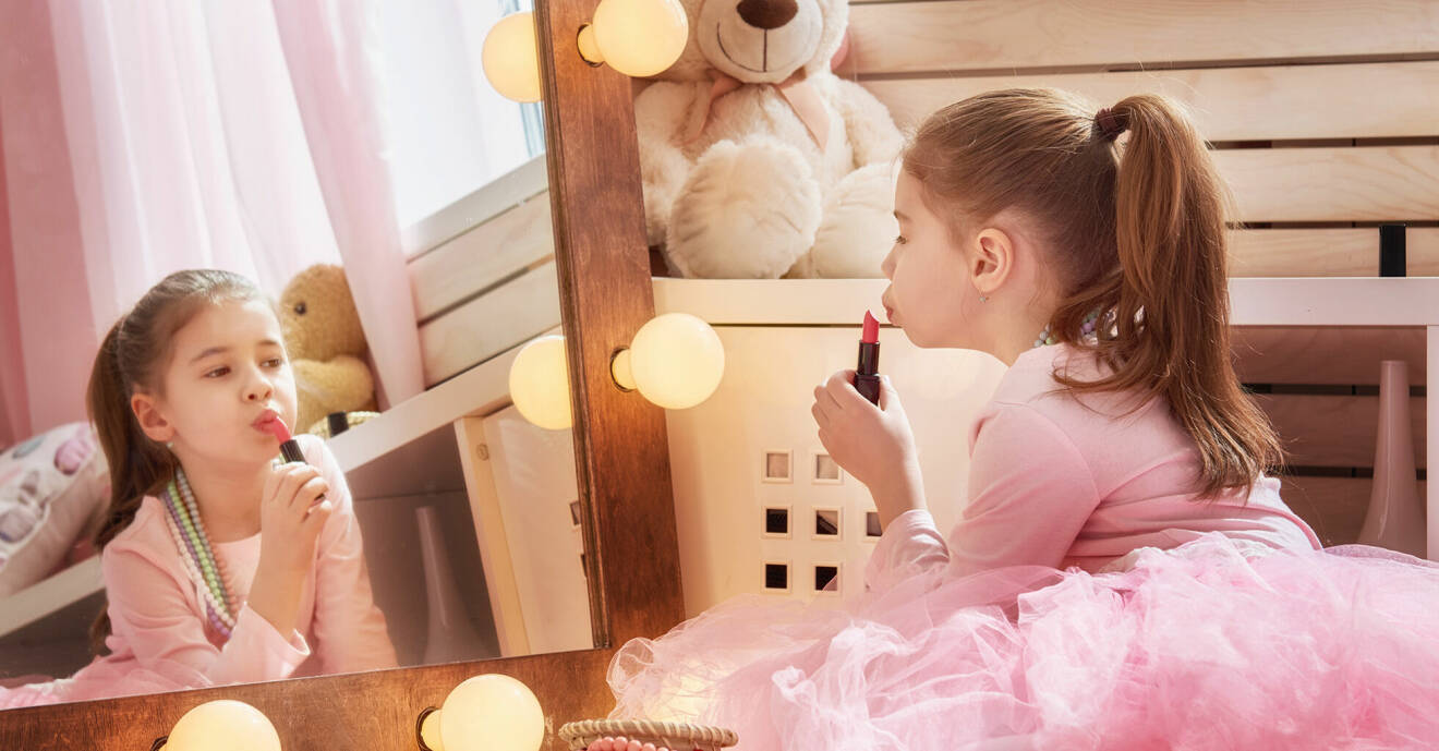 Brunhårig flicka i rosa ballerinaklänning sitter framför spegeln och målar läppstift på läpparna.
