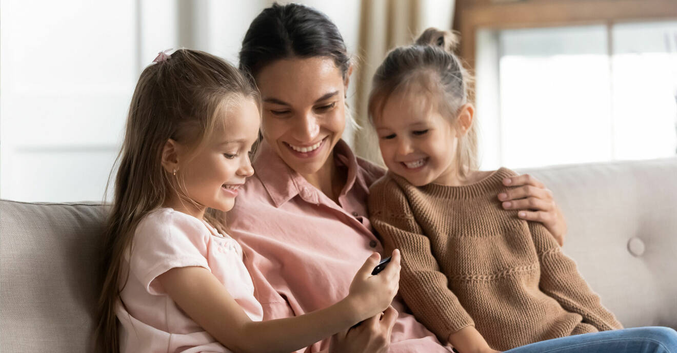 Vuxen kvinna sitter mellan sina två unga döttrar i en soffa. De kollar på en telefon och skrattar.