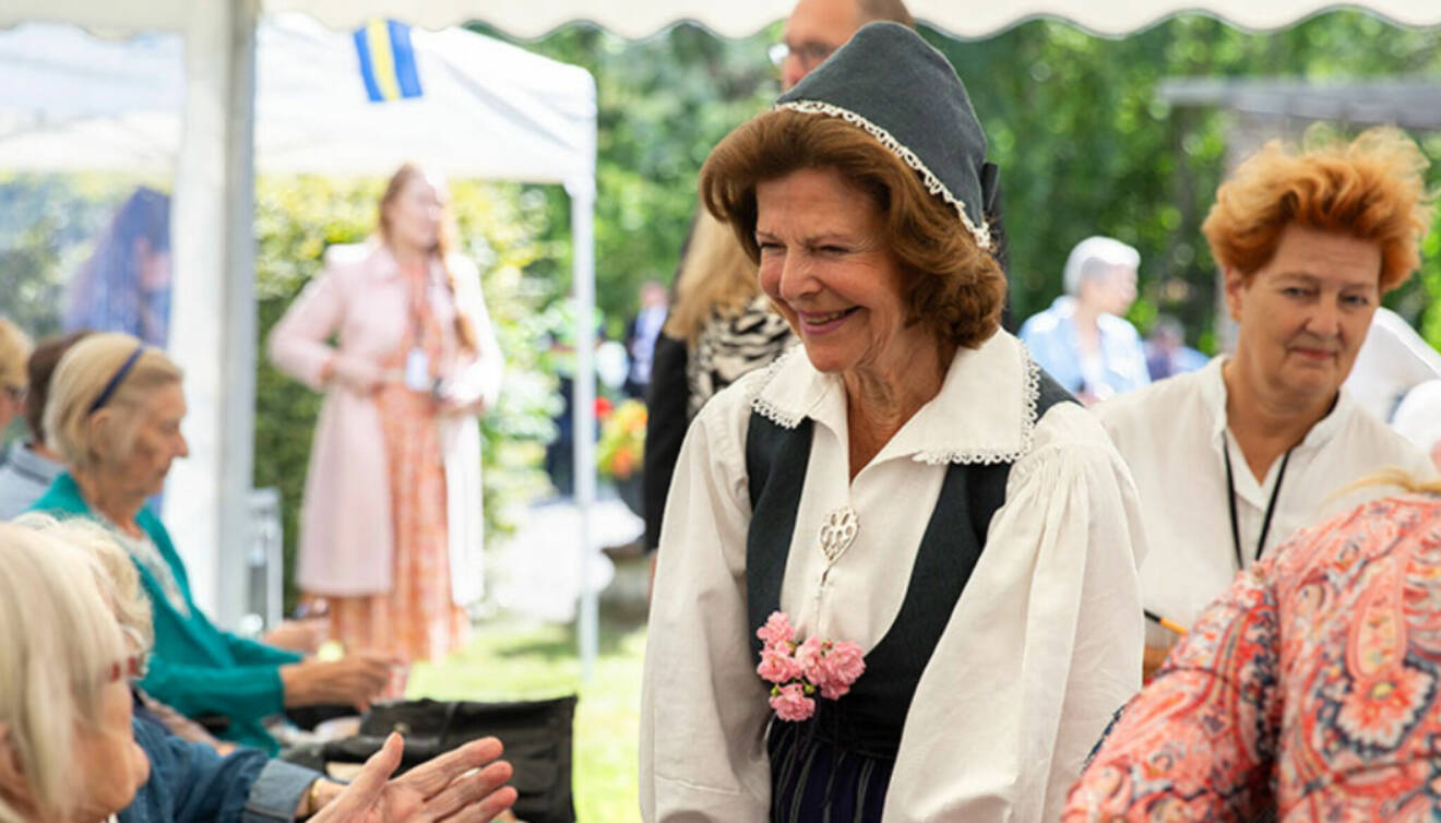 Drottning Silvia i folkdräkt talar med en äldre kvinna på De gamlas dag på Ekerö 2022, bakom henne står författaren Ingrid Thörnqvist.