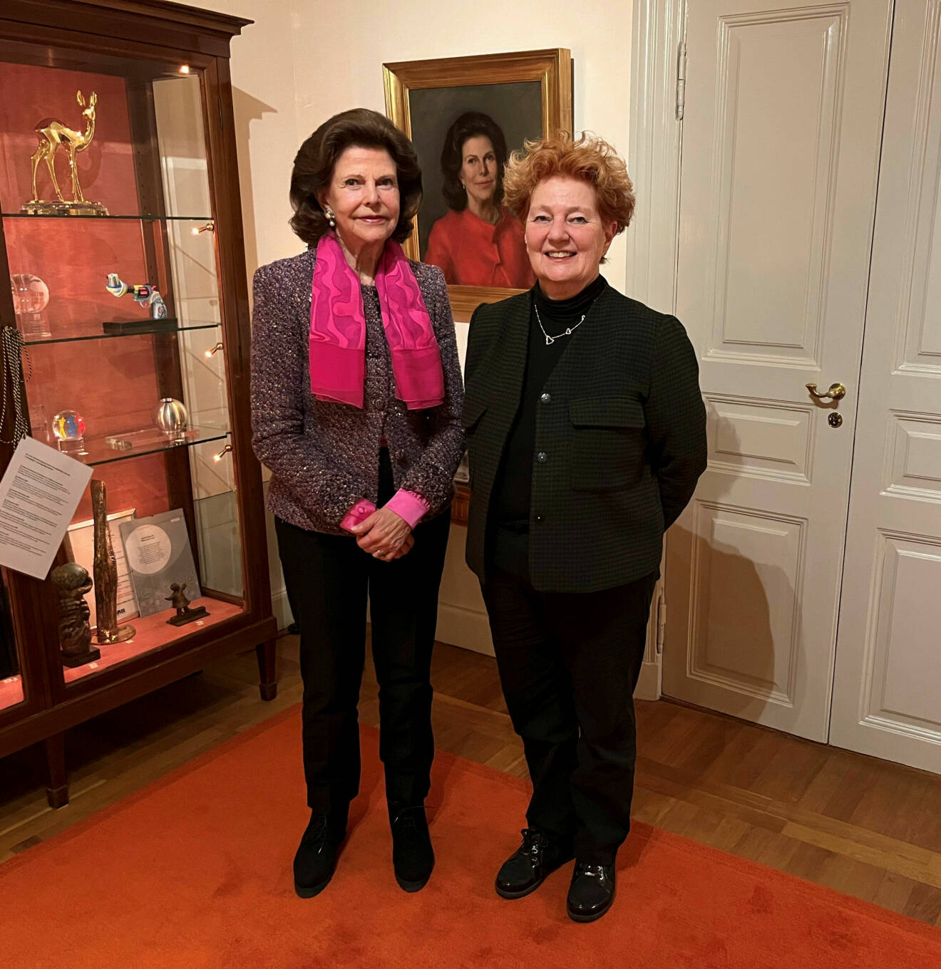 Drottning Silvia tillsammans med författaren Ingrid Thörnqvist fotograferade på Slottet, framför ett målat porträtt av drottningen.