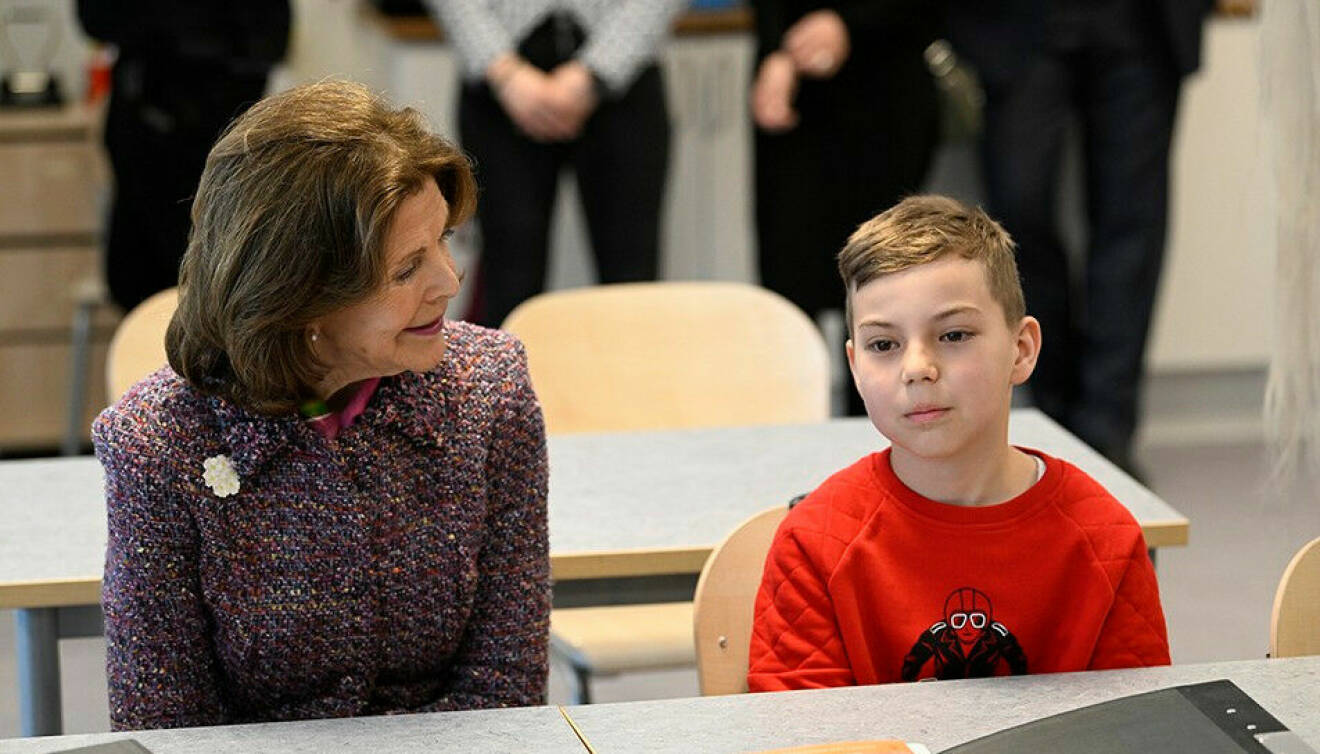 Drottning Silvia sitter i en skolbänk sida vid sida med en liten ukrainsk pojke på ungefär 10 år i röd tröja.