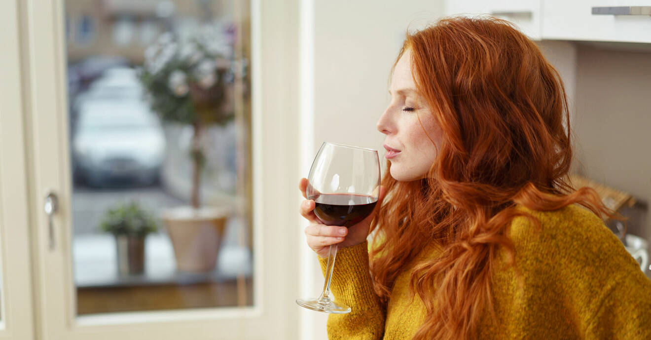 Rödhårig kvinna i 30-årsåldern dricker ett glas rött vin inomhus.