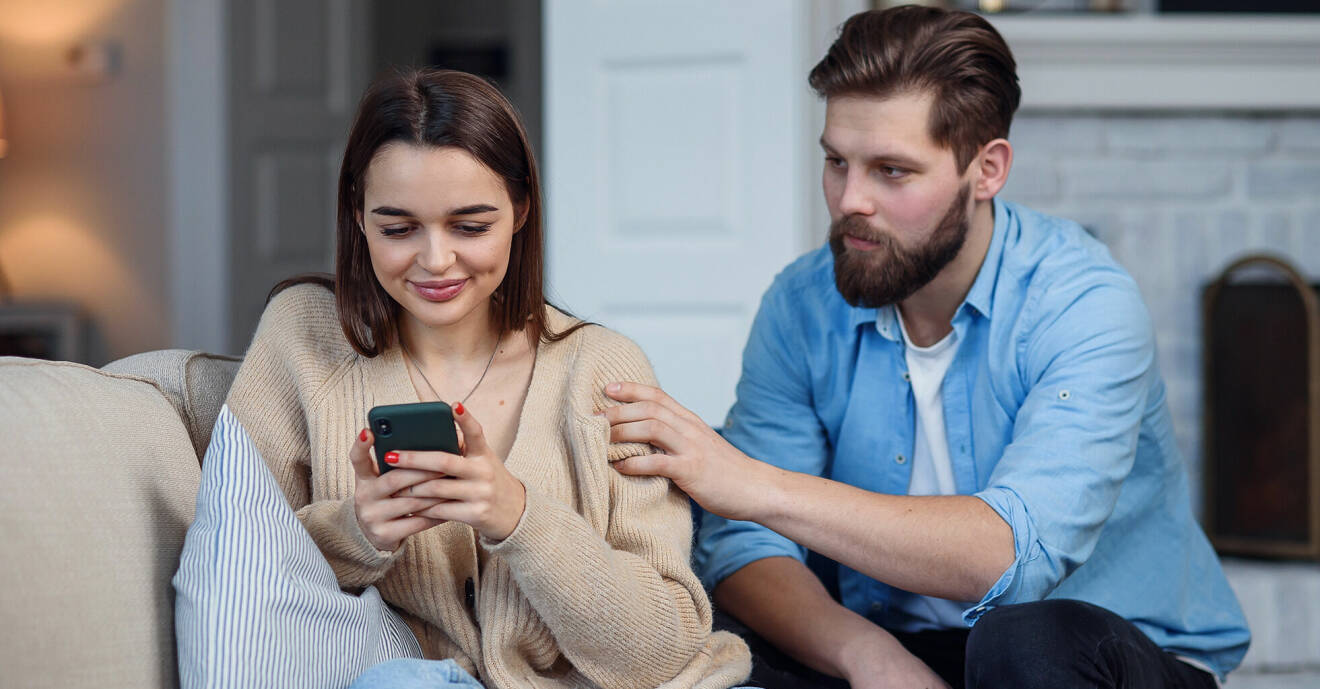 Kvinna i 30-årsåldern sitter i soffan och kollar ner i sin mobiltelefon. Bredvid henne sitter en man i samma ålder och försöker få hennes uppmärksamhet.