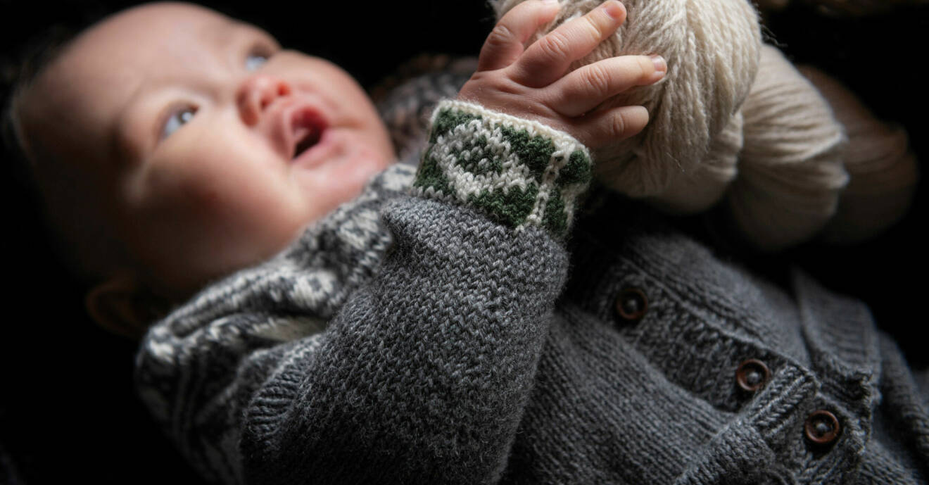 En bebis har på sig en grå stickad mönstrad kofta och hen håller i ett vitt garnnystan.
