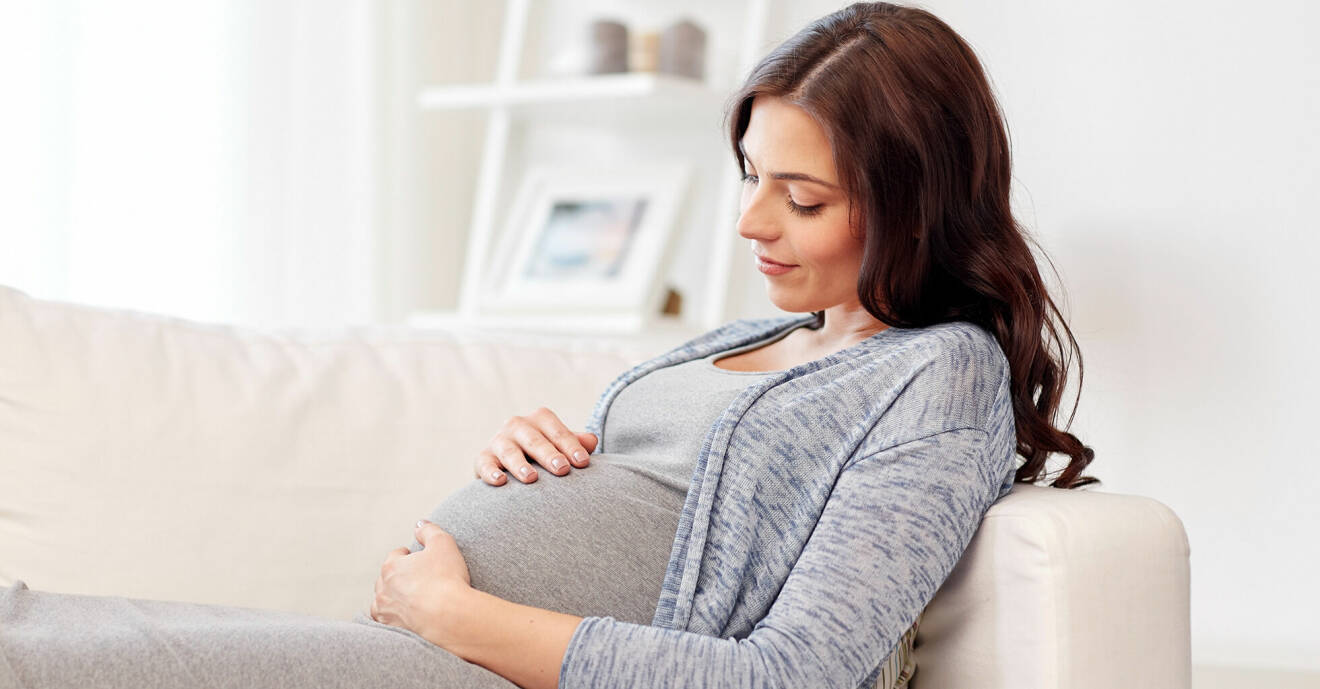 Gravid kvinna i 30-årsåldern sitter i en soffa och håller sina händer på sin mage.