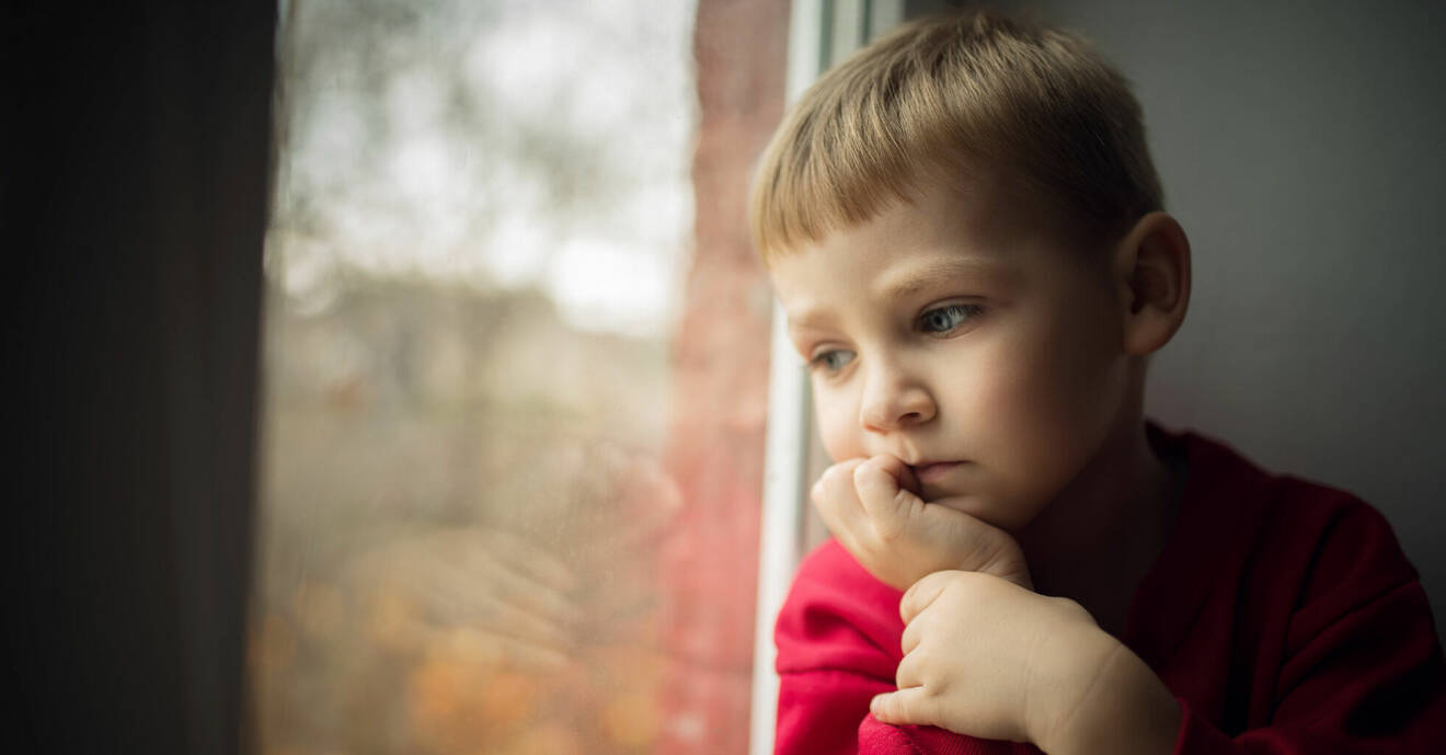 Liten pojke sitter och tittar ut genom ett fönster. Ser ledsen ut.