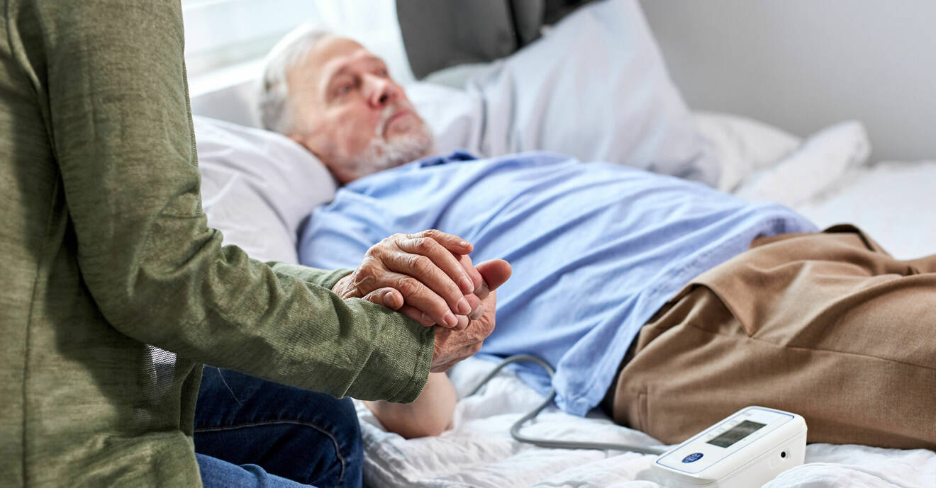 Äldre man ligger i en säng och håller i en kvinnas hand.