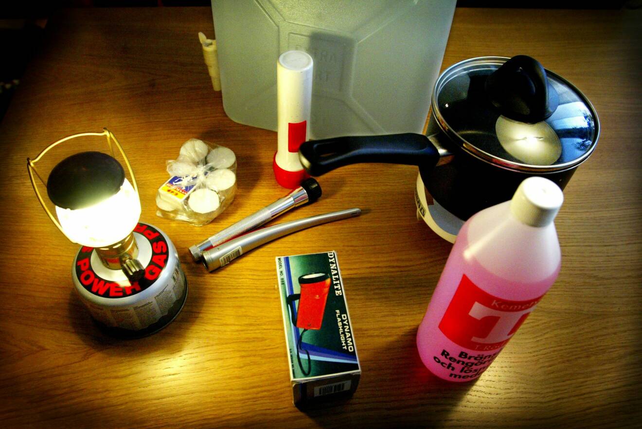 Gasdriven lampa, värmeljus, ficklampor, nödradio, vattendunk och T-röd på ett bord hos en prepper.