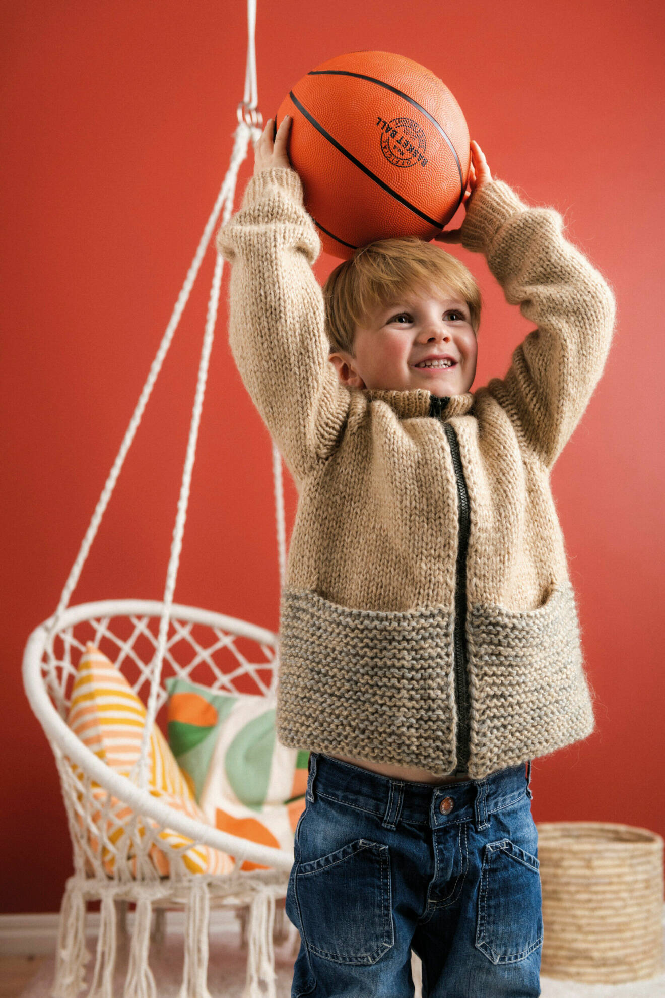 Pojke har på sig en beige stickad tröja och håller i en basketboll med armarna uppsträckta över huvudet.