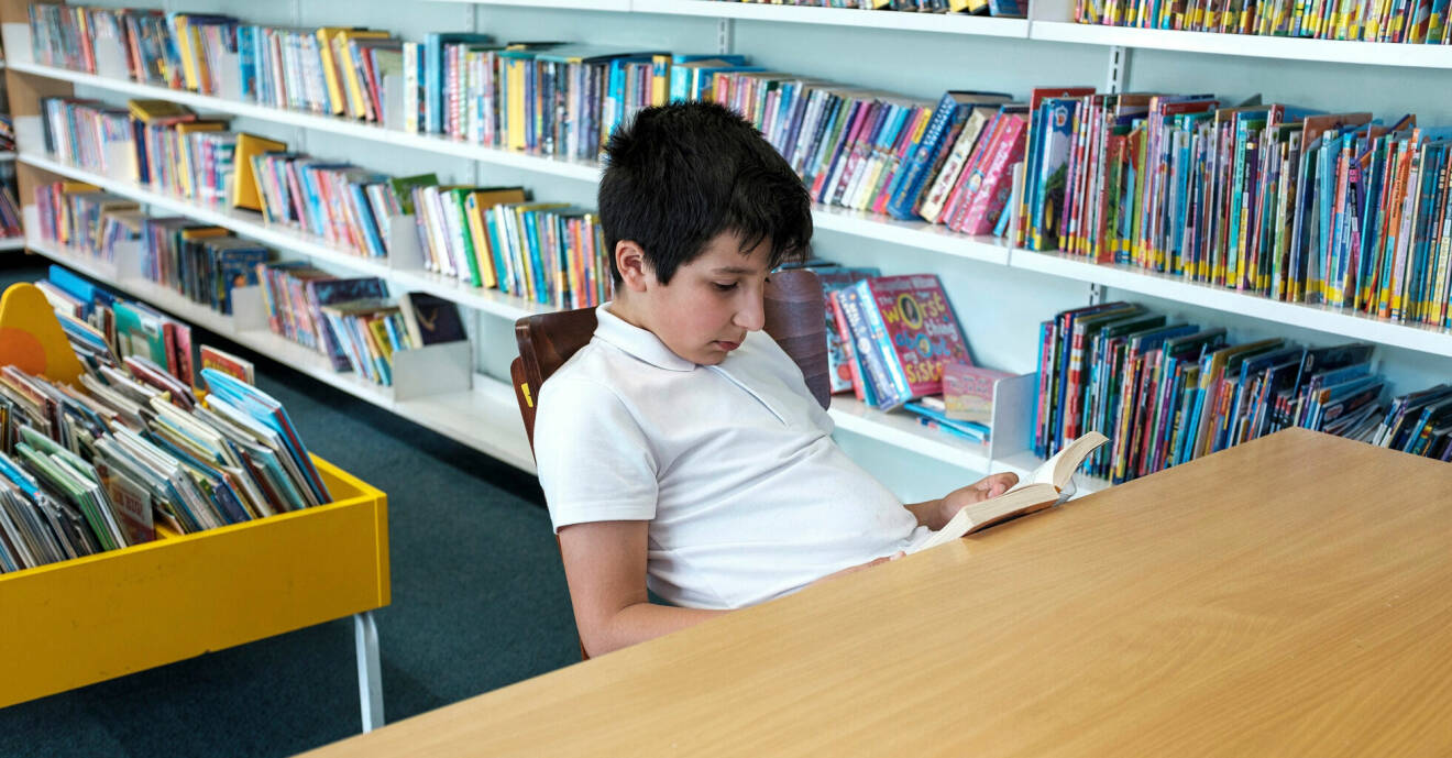 Ung kille sitter i ett bibliotek och läser en bok.