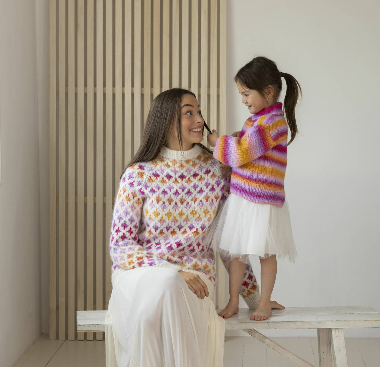 Kvinna och liten flicka har på sig färgglada stickad tröjor och vita kjolar.