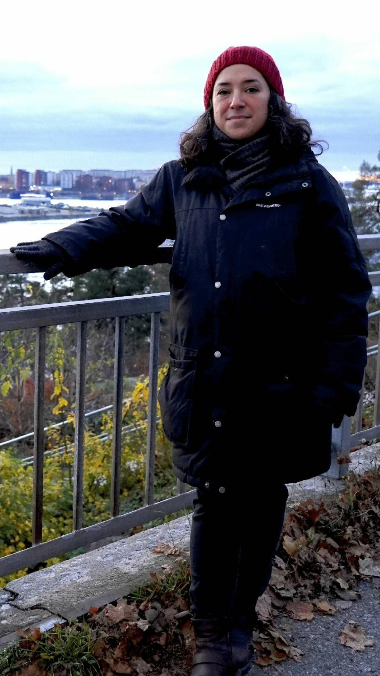 En mörkhårig kvinna står vid ett räcke med utsikt över Stockholm. Hon bär en svart jacka av märket Everest och har en röd mössa på sig.