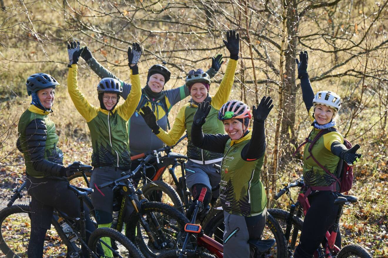 Sex kvinnor på varsina mountainbikes. De ler mot kameran och höjer sina händer i luften.