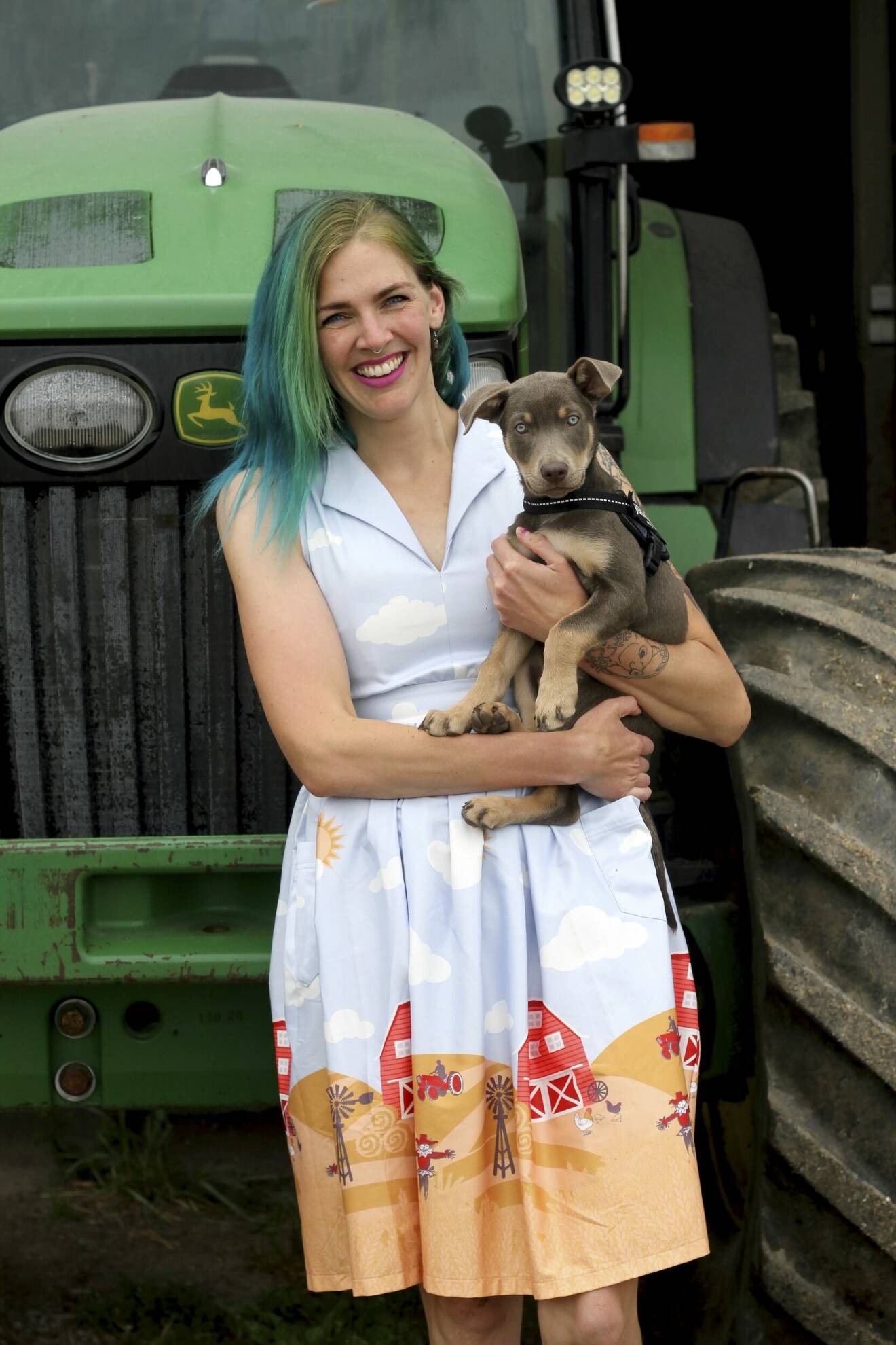 Emilia Astrenius har grönt hår och står och håller i en hundvalp. Hon bär klänning med bonde-tema och står framför en grön traktor.