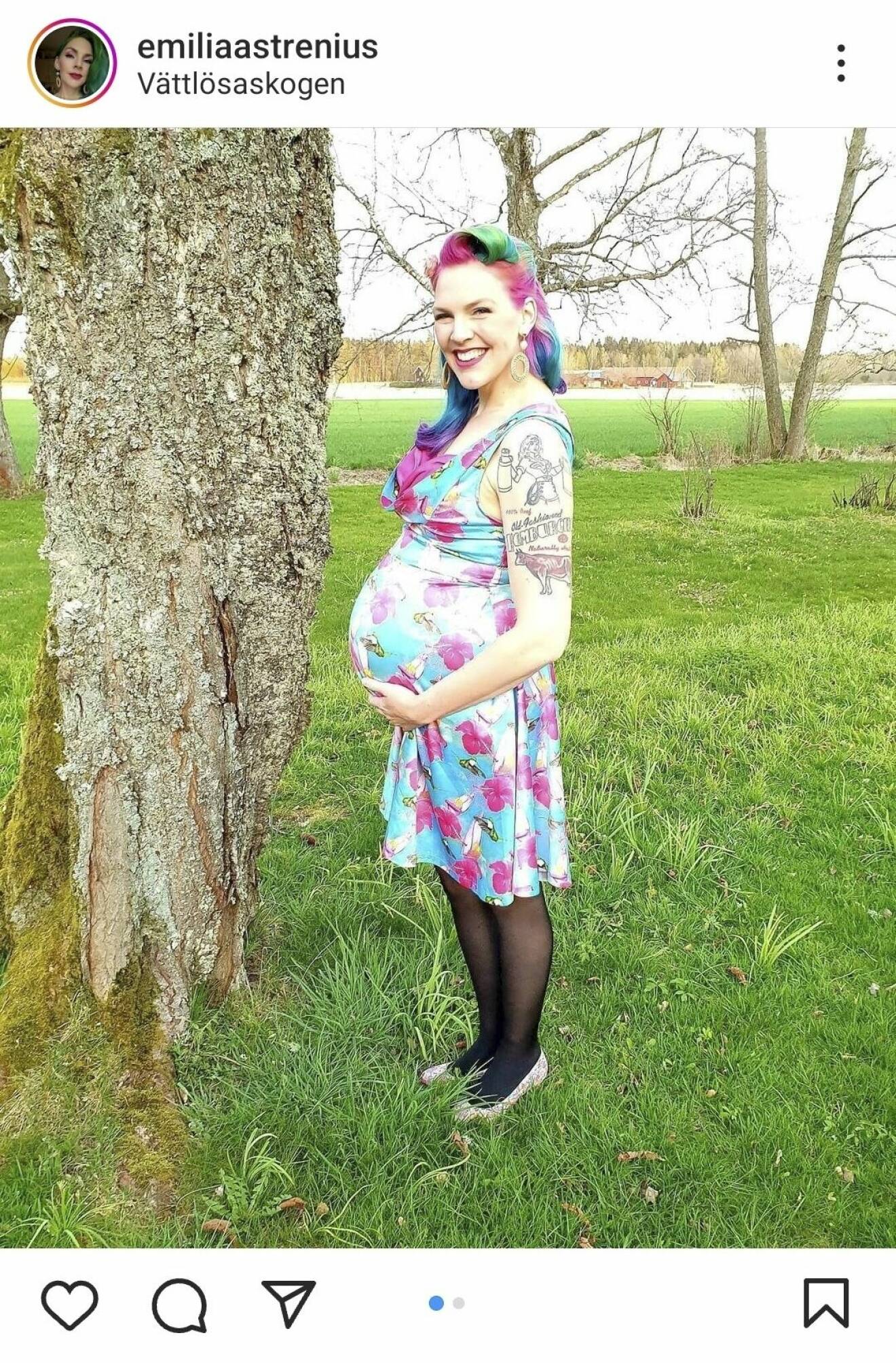 Emilia Astrenius är gravid och ler mot kameran. Hon håller sig på magen och bär en färgglad klänning. Även hennes hår är färgglatt.