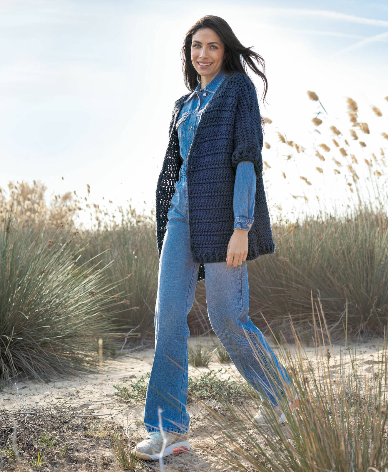 Kvinna står på en strand med jeans och en blå virkad kofta.