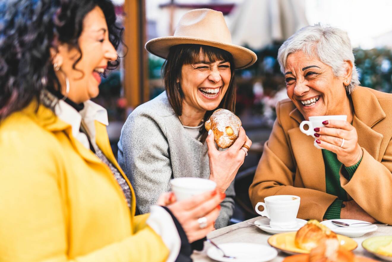 Tre kvinnor skrattar och dricker kaffe tillsammans.