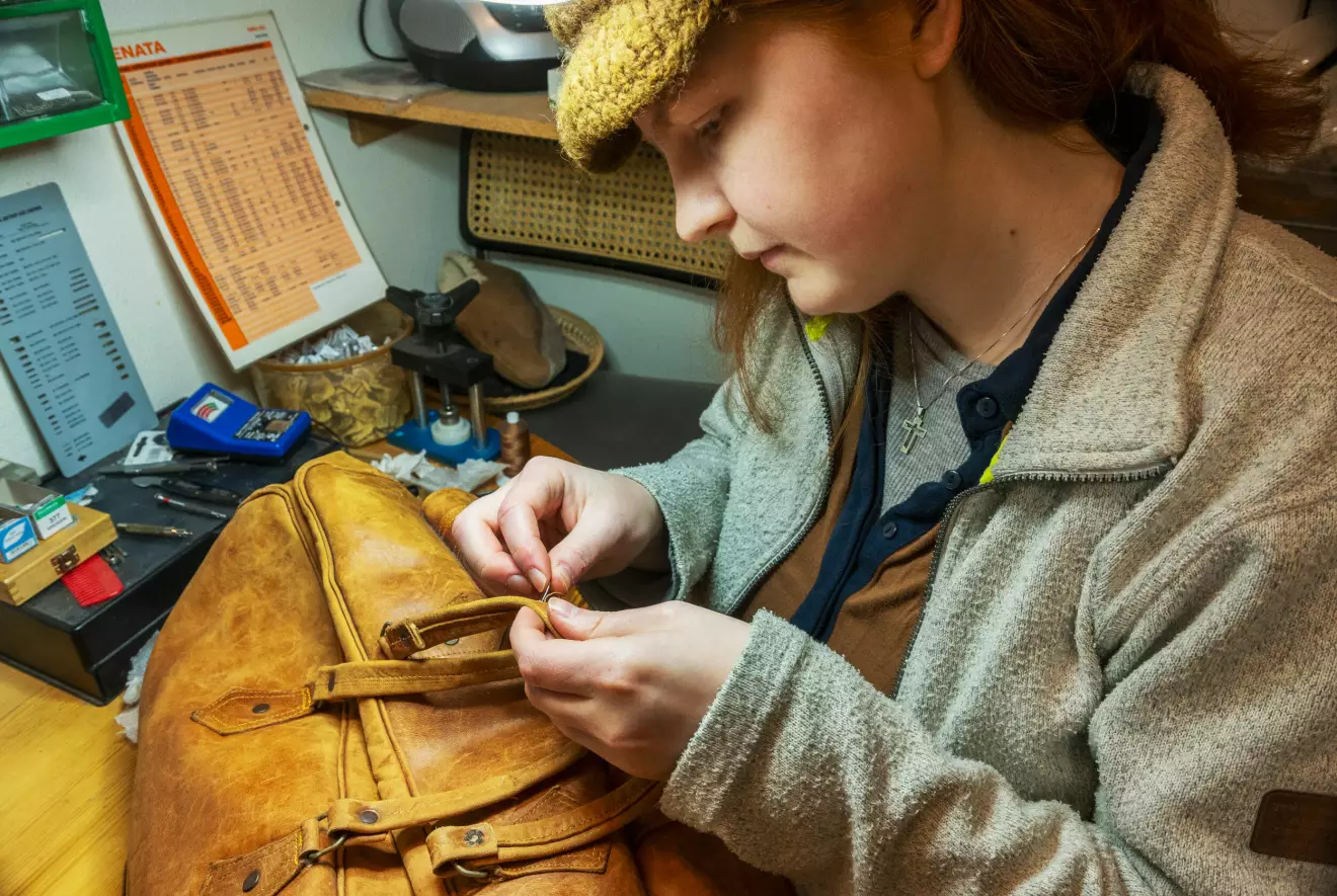 Lisabet syr i handtag på gul-brun läderväska vid sitt arbetsbord.