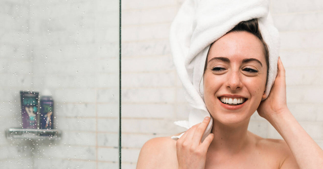 En kvinna som ler i duschen med en handduk på huvudet.