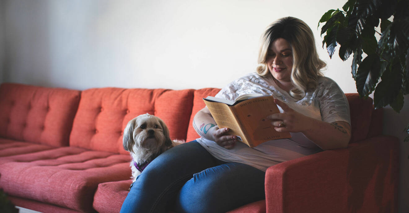 Kvinna sitter i soffan och läser en bok med hund som sällskap.