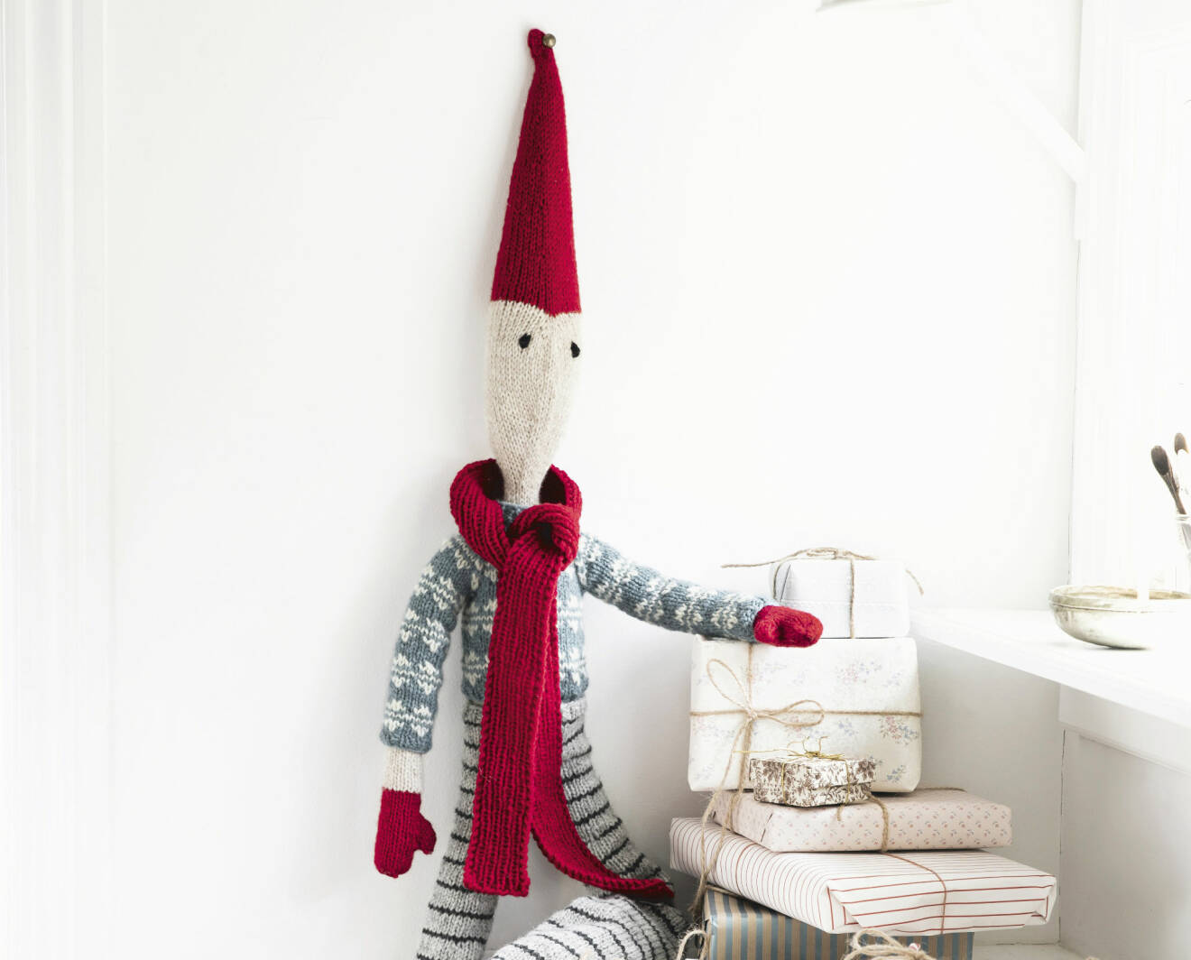 Detaljbild av stickad jultomte som är fäst på väggen och håller armen på en hög julklappar.